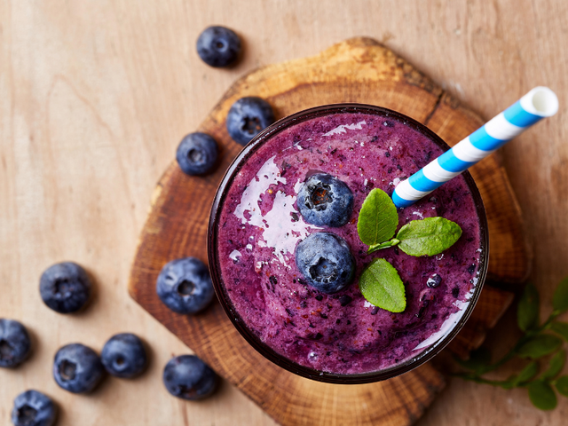 Top view of a blueberry blast Complete shake in a glass with a striped straw, garnished with fresh blueberries and mint, placed on a wooden board with scattered blueberries around.