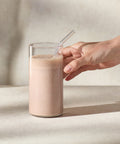 Glass of a Juice Plus+ Complete Chocolate shake with a straw, held by a hand on a neutral background