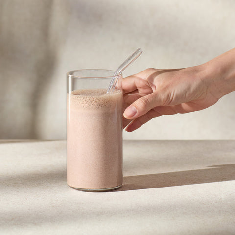 Glass of a Juice Plus+ Complete Chocolate shake with a straw, held by a hand on a neutral background