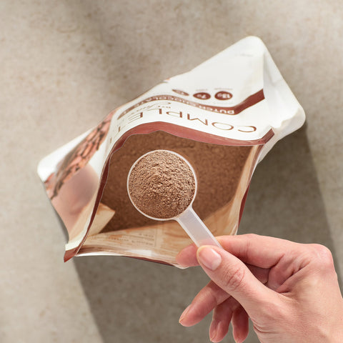 Hand holding a package of Juice Plus+ Chocolate powder with a spoonful of powder visible.