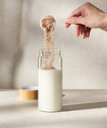 Person pouring a scoop of Juice Plus+ Complete powder into a glass of milk on a neutral background