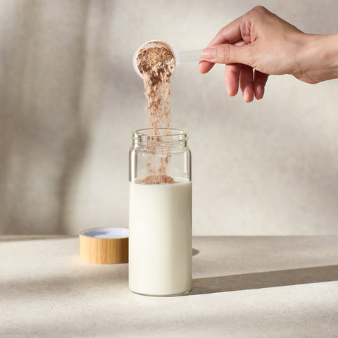 Person pouring a scoop of Juice Plus+ Complete powder into a glass of milk on a neutral background