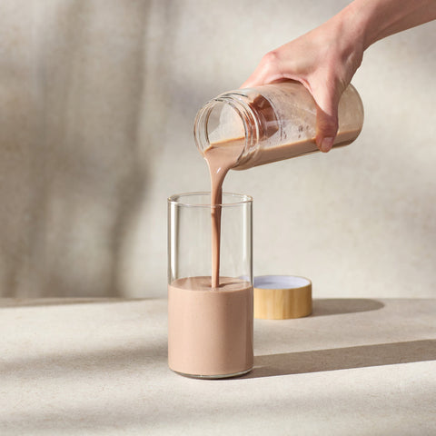 Person pouring a creamy liquid from a glass jar into another glass on a neutral background