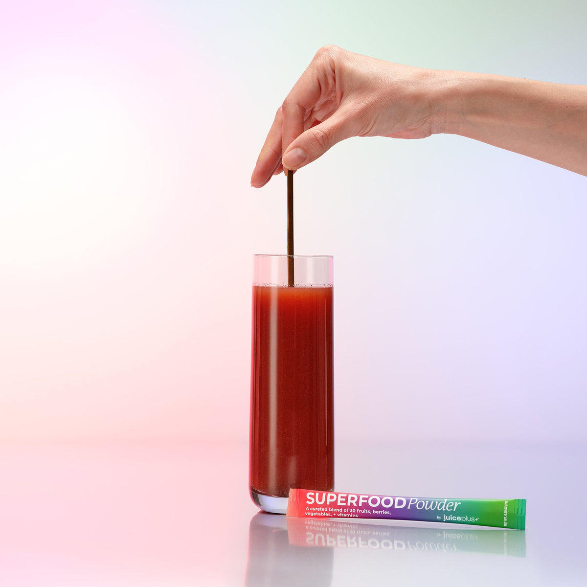 Hand stirring a glass of Juice Plus+ Superfood Powder package on a gradient background