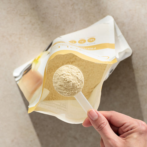 Hand holding a spoon with Juice Plus+  Complete powder over a package of the same powder on a neutral background