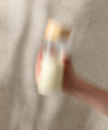 Blurry image of a hand holding a small bottle with a gold cap against a neutral background