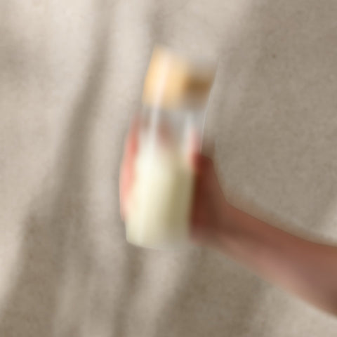 Blurry image of a hand holding a small bottle with a gold cap against a neutral background