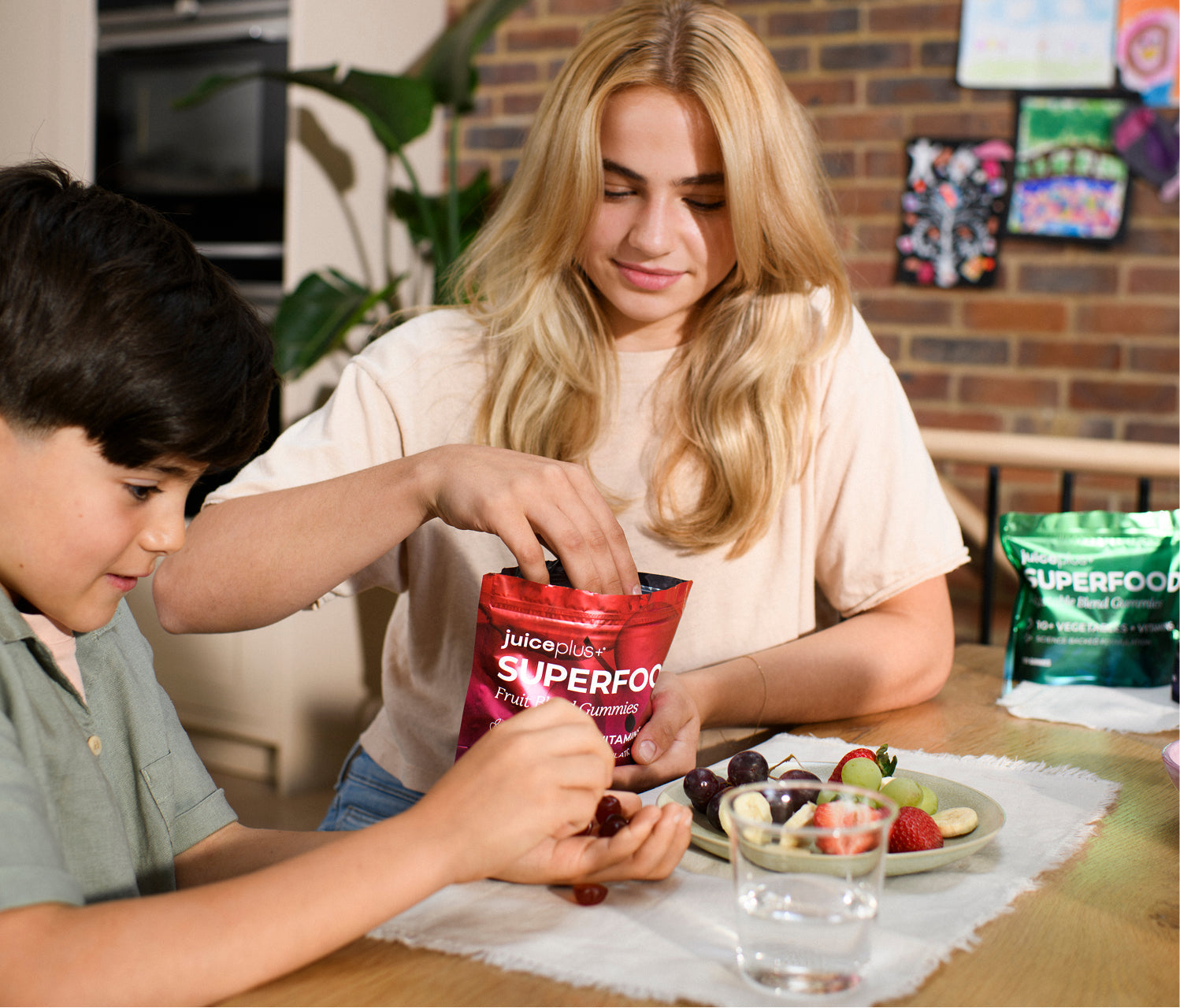 A lifestyle scene showing a young boy and a woman sitting at a wooden table, with the woman opening a red bag of Juiceplus+ Superfood powder. A green bag of Juiceplus+ Superfood is visible on the table to the right, along with a plate of fresh fruit and a glass of water.