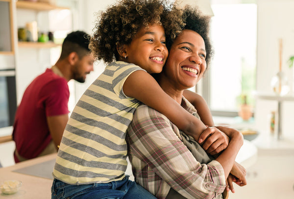 A smiling woman carries a young child on her shoulders in a bright kitchen, with another man blurred in the background.