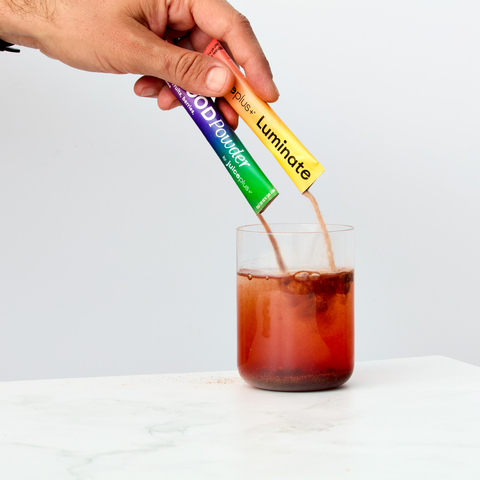 Hand pouring a Juice Plus+ Luminate and Superfood Powder into a glass of iced coffee on a white background