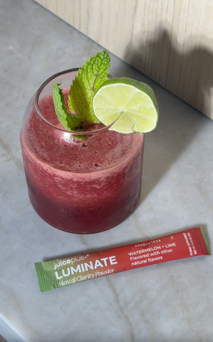 A glass of pinkish-red beverage garnished with mint leaves and a lime slice, placed on a light marble-like surface next to a Juice Plus+ Luminate vertical clarity powder stick pack.