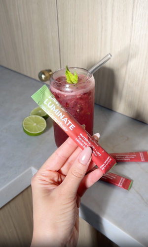 A hand holds a red and green Luminate oral supplement stick in front of a glass of pink drink on a kitchen counter.